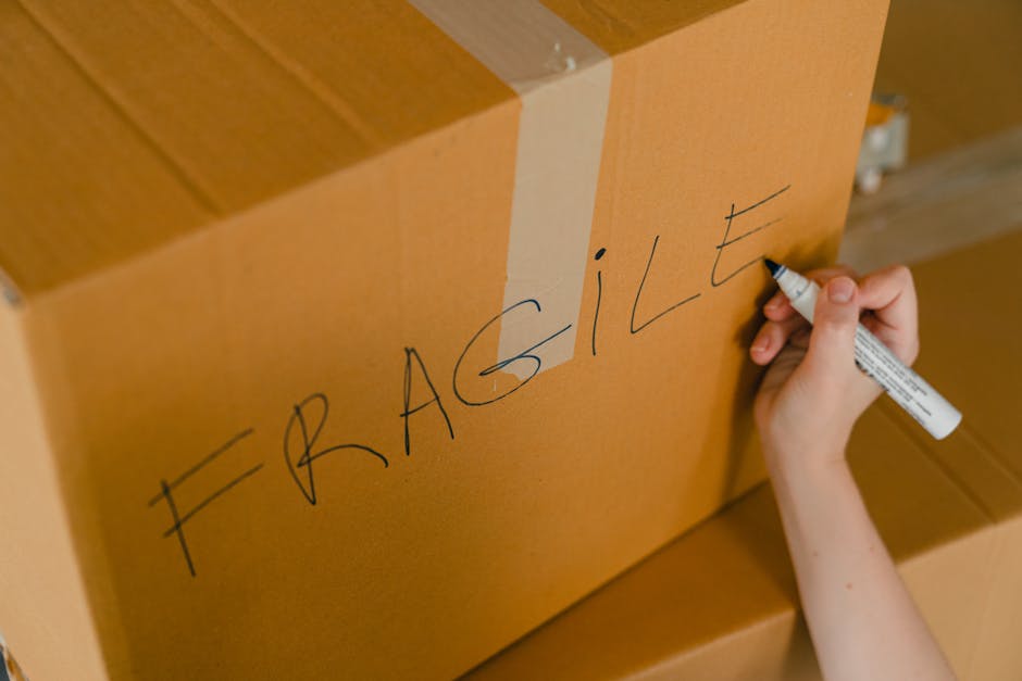 A person sitting cross-legged on a wooden floor inside a home, wrapping fragile glassware with protective packing materials. They are holding a roll of clear packing tape and carefully applying it to