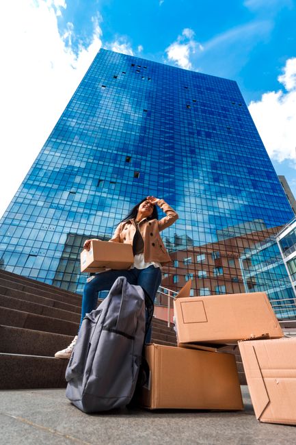 A woman sitting on outdoor steps in front of a modern glass office building, surrounded by packed moving boxes made of cardboard and she is holding a smaller box in her arms. Nearby, there is a large