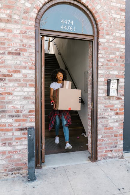 A woman sitting on outdoor steps in front of a modern glass office building, surrounded by packed moving boxes made of cardboard and she is holding a smaller box in her arms. Nearby, there is a large